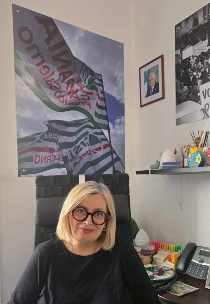 Ritratto di una donna con occhiali seduta a una scrivania, con bandiere sindacali sullo sfondo nella stanza.