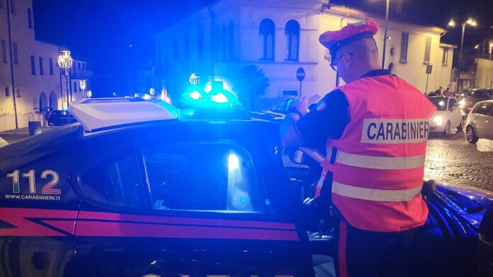 Carabiniere in divisa con giubbotto catarifrangente davanti auto dei Carabinieri con lampeggianti accesi durante controllo notturno a Solofra