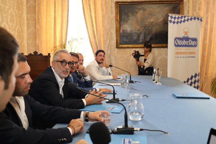 Sala con tavolo blu durante la conferenza stampa di presentazione di OktoberDays a Napoli, con partecipanti seduti e pubblico in ascolto.