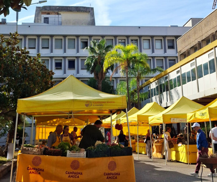 Stand gialli allestiti all'aperto con persone e prodotti alimentari, in un cortile ospedaliero con palazzi sullo sfondo.