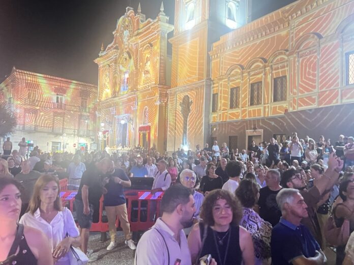 Una piazza affollata di persone di sera con videomapping colorato sulle facciate degli edifici storici, foto illustrativa della festa patronale a Frattamaggiore.