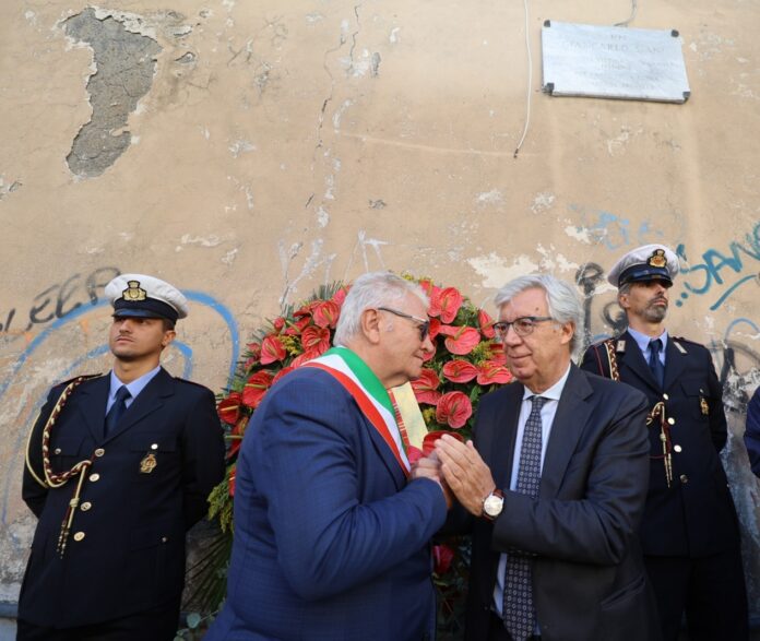 Cerimonia con persone in piedi vicino a una corona di fiori rossi contro un muro con targa commemorativa.