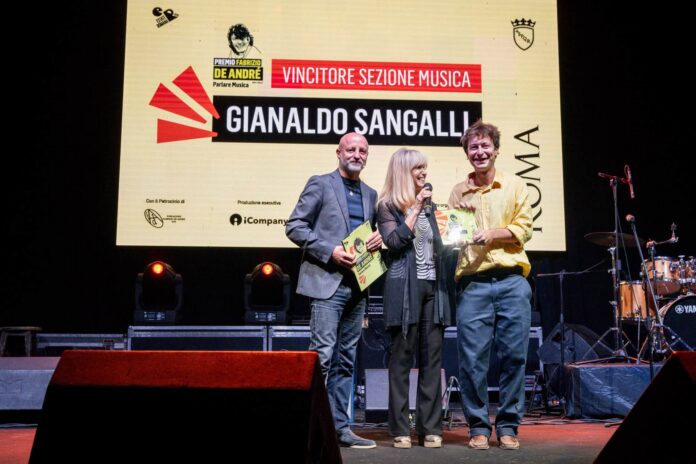 Premiazione dei vincitori della sezione musica del Premio Fabrizio De André sul palco con sfondo schermo digitale.