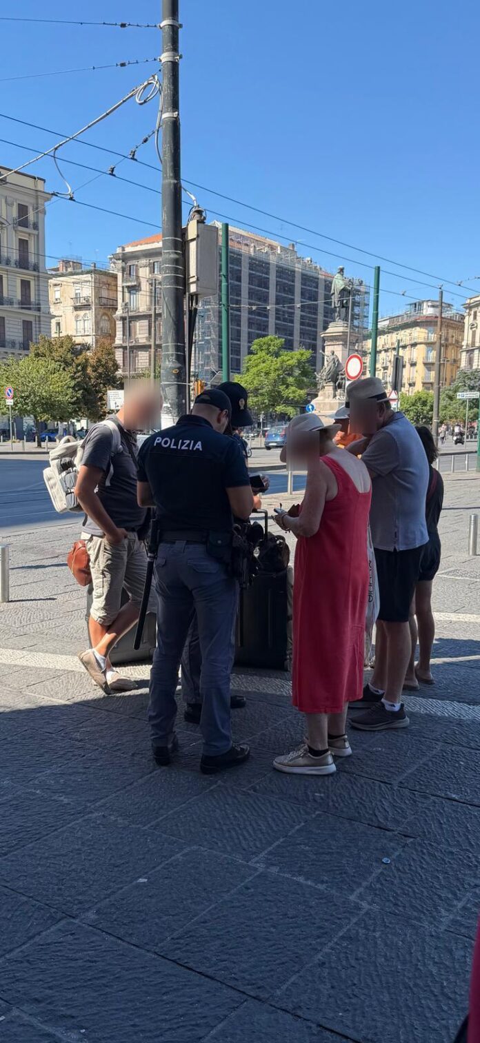 Turisti e agente di polizia in piazza Garibaldi a Napoli, con sfondo di edifici e traffico urbano.