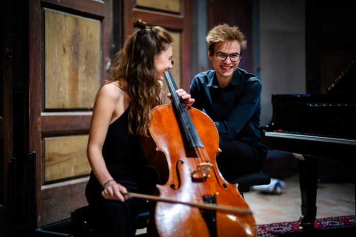 Giovani musicisti con violoncello e pianoforte durante una prova o una performance in una sala storica.