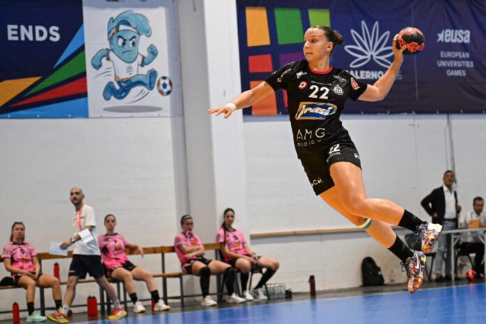 Giocatrice di pallamano della Jomi Salerno in salto con la palla in mano durante una partita su campo coperto, con compagne di squadra sedute sulla panchina sullo sfondo.