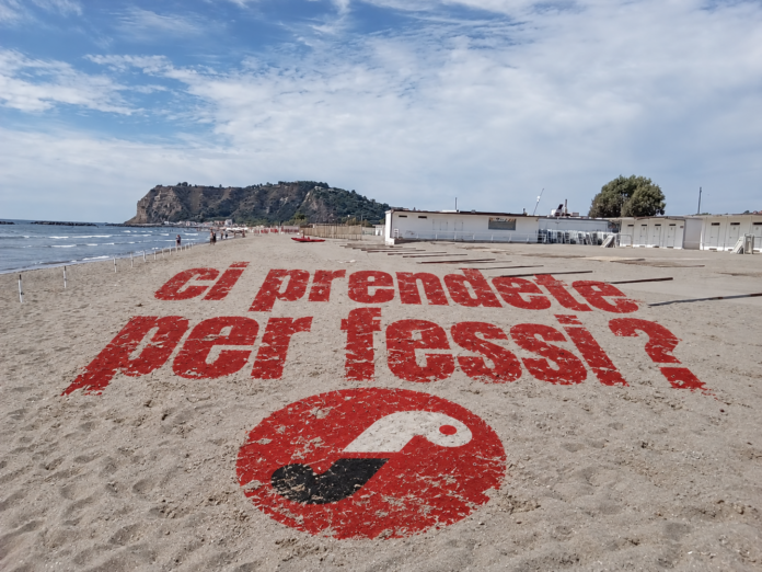 Spiaggia di Bacoli con messaggio scritto sulla sabbia 'ci prendete per fessi?' in caratteri rossi.
