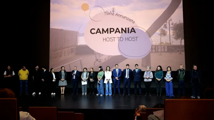 Un gruppo di persone è disposto sul palco di una sala; alle loro spalle compare la scritta «Campania Host to Host».
