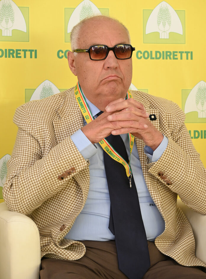 Foto di un uomo anziano con occhiali scuri in giacca beige, seduto davanti a uno sfondo con logo Coldiretti.