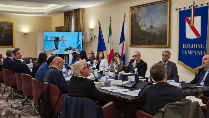 Persone sedute attorno a un tavolo di riunione; sullo sfondo sono visibili bandiere e un simbolo della Regione Campania.
