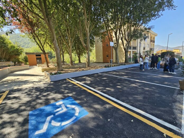 Area parcheggio con stalli riservati a persone con disabilità e gruppi di persone sullo sfondo in una giornata di sole a Fusara.