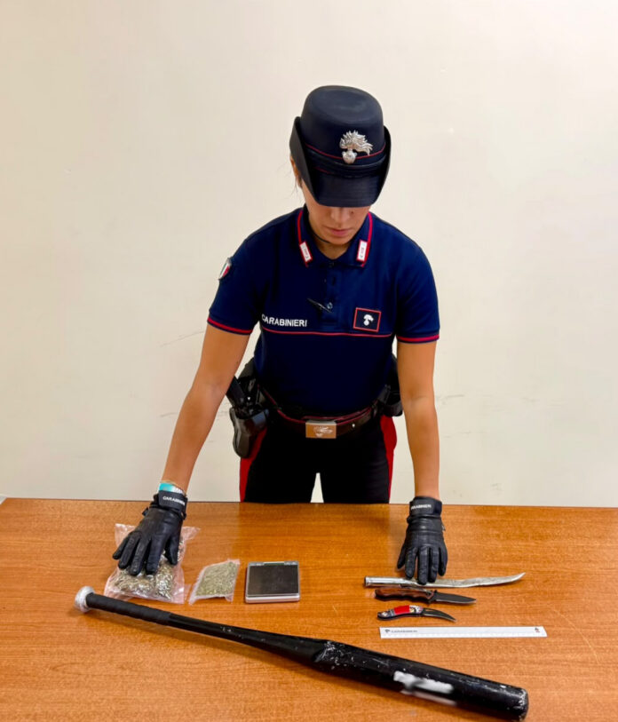 Carabiniere davanti a un tavolo con coltelli, mazza da baseball, bilancino e droga sequestrata.