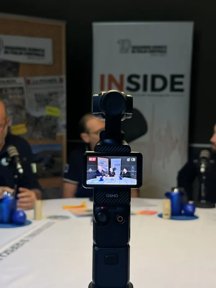 Una videocamera è in primo piano; sullo sfondo si vedono persone sedute a un tavolo e un pannello con la scritta “INSIDE”.
