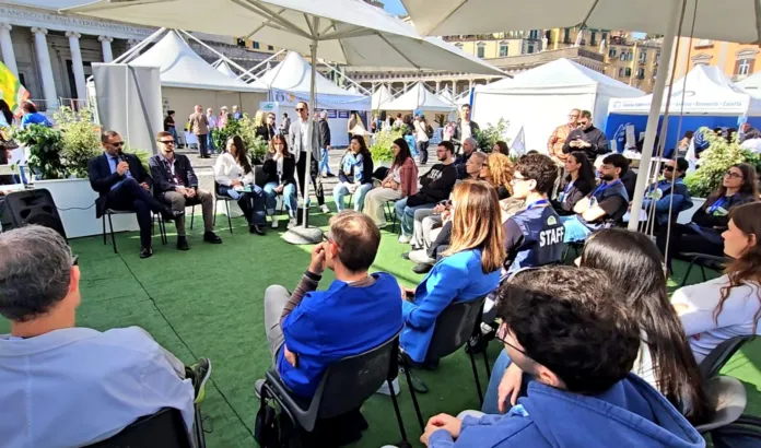 Persone sedute e in piedi ascoltano un incontro all’aperto, sotto gazebo.