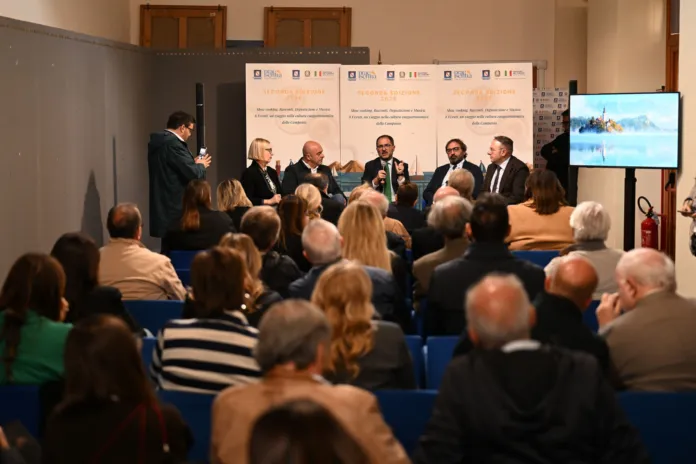 Sala con persone sedute in platea e un gruppo di relatori su sedie davanti a pannelli espositivi.