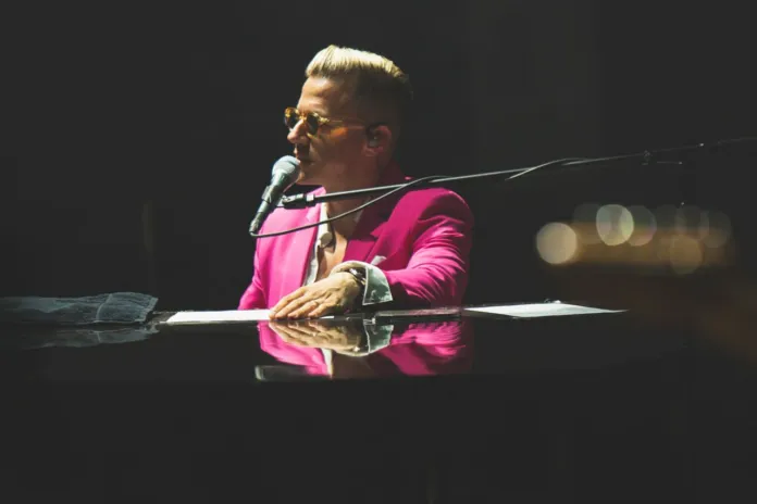 Un uomo con una giacca fucsia suona al pianoforte e canta al microfono, sotto luci di scena.