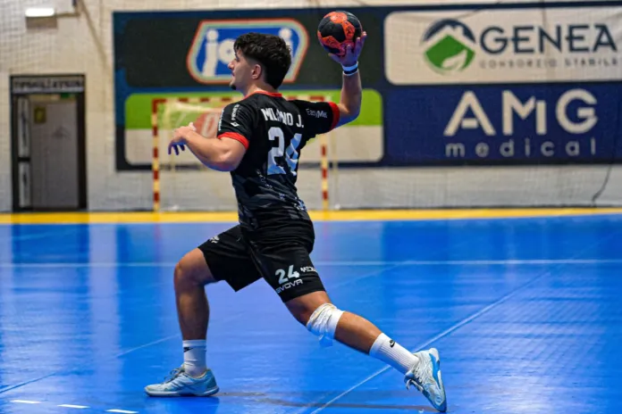 Un giocatore con maglia nera avanza sul campo da pallamano e solleva il pallone per lanciare.