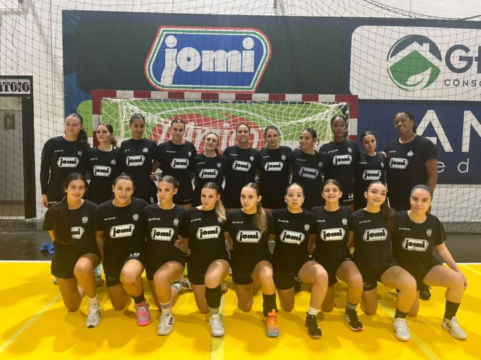 Squadra femminile della Jomi Salerno in posa in palestra davanti alla porta di pallamano, indossano magliette nere con logo jomi.