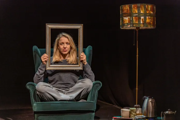 Immagine di scena teatrale con attrice Viola Graziosi che impersona un personaggio seduta su una poltrona con cornice, attorno oggetti di scena, sfondo scuro, illuminazione concentrata sulla protagonista.