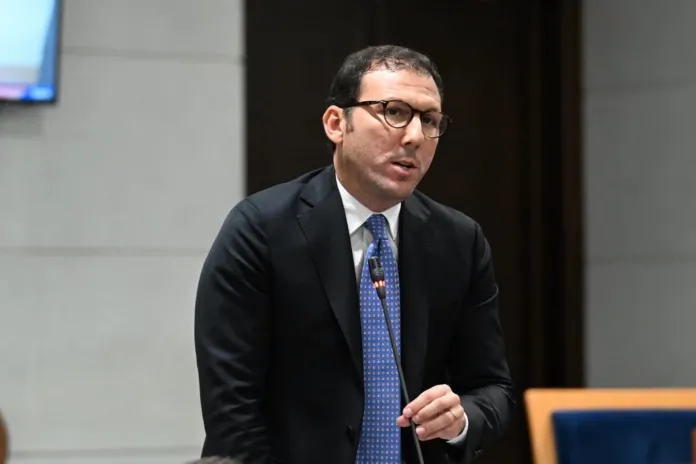 Uomo con occhiali e abito formale che parla in pubblico davanti a un microfono.