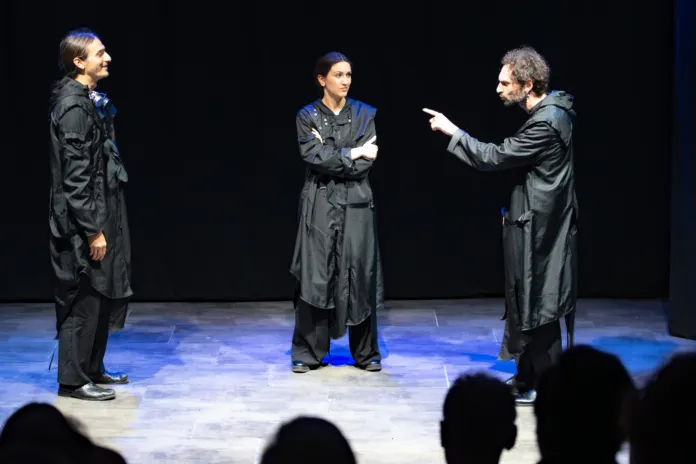 Tre persone in costume scuro su un palcoscenico: una indica un’altra, mentre la terza osserva con le braccia conserte.