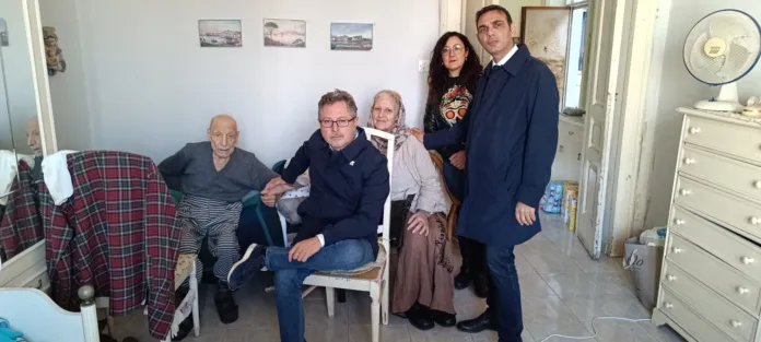 In una stanza, due uomini anziani sono seduti accanto a un uomo; dietro di loro ci sono una donna e un altro uomo in piedi.