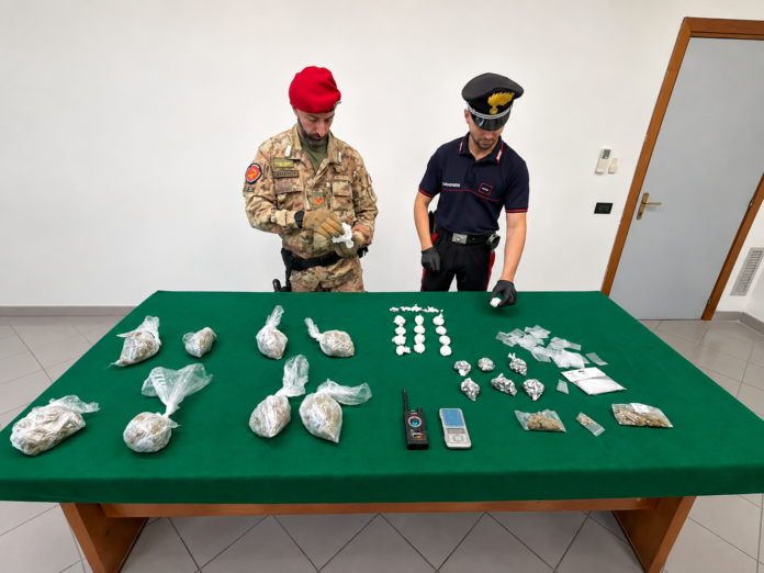 Due carabinieri in uniforme controllano dosi di droga, un bilancino e un rilevatore di microspie disposti su un tavolo verde.