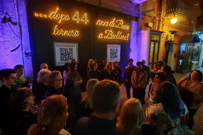 Immagine di un evento culturale con pubblico presente di sera, vicino a un edificio illuminato, atmosfera calda e vivace.