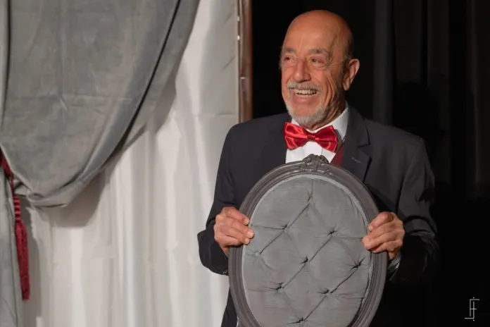 Immagine di un attore su palco durante una rappresentazione teatrale, con un buffo orologio a forma di coppa davanti a lui, che sorride mentre interpreta un personaggio comico