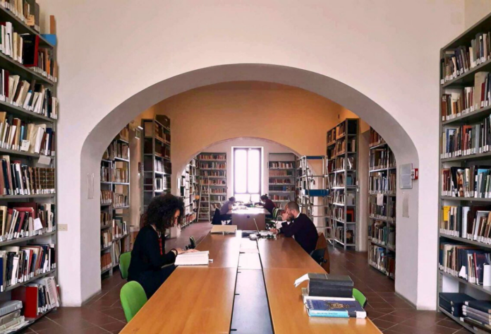 Interno di una biblioteca con persone sedute a un tavolo che leggono, scaffali pieni di libri ai lati.
