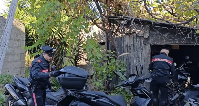 Due persone in uniforme si trovano accanto a scooter davanti a un capanno rurale.