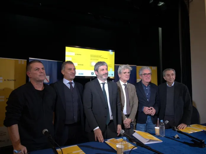 Sei persone in abiti formali dietro un tavolo con cartelle e bottiglie d’acqua; alle loro spalle si vede uno schermo.