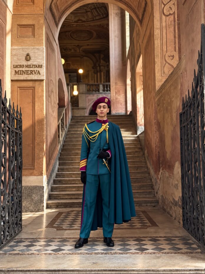 Attore in costume militare con cappello e mantello davanti a un ingresso decorato con scritta 'Liceo Militare Minerva'.