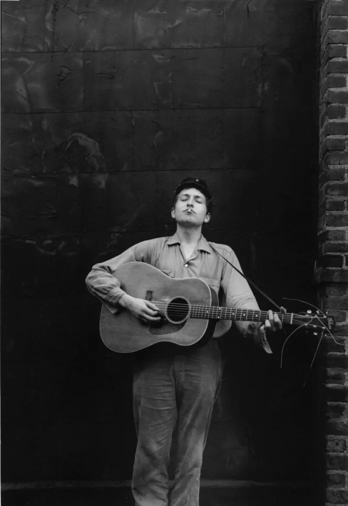 Ritratto in bianco e nero di Bob Dylan con una chitarra acustica, davanti a una parete di mattoni.