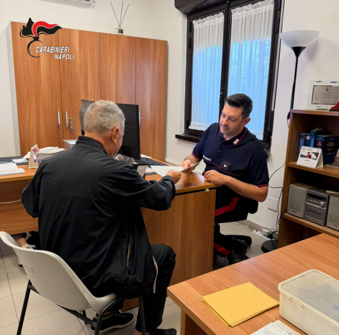 Un carabiniere al computer in ufficio mentre parla con un uomo seduto di fronte a lui.