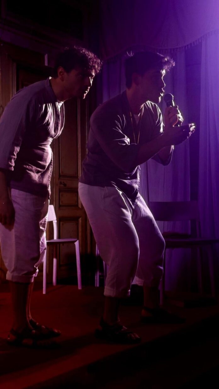 Attori in scena durante uno spettacolo teatrale con luci di scena violacee, in interno.