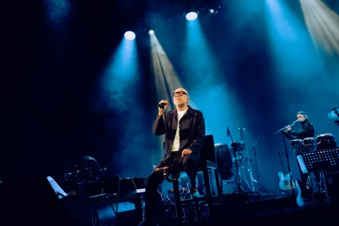 Un cantante con microfono è seduto al centro di un palco illuminato; un musicista suona a destra.