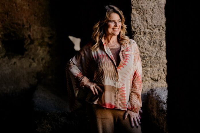 Monica Sarnelli sorridente con una camicia colorata in un ambiente interno con parete di pietra.
