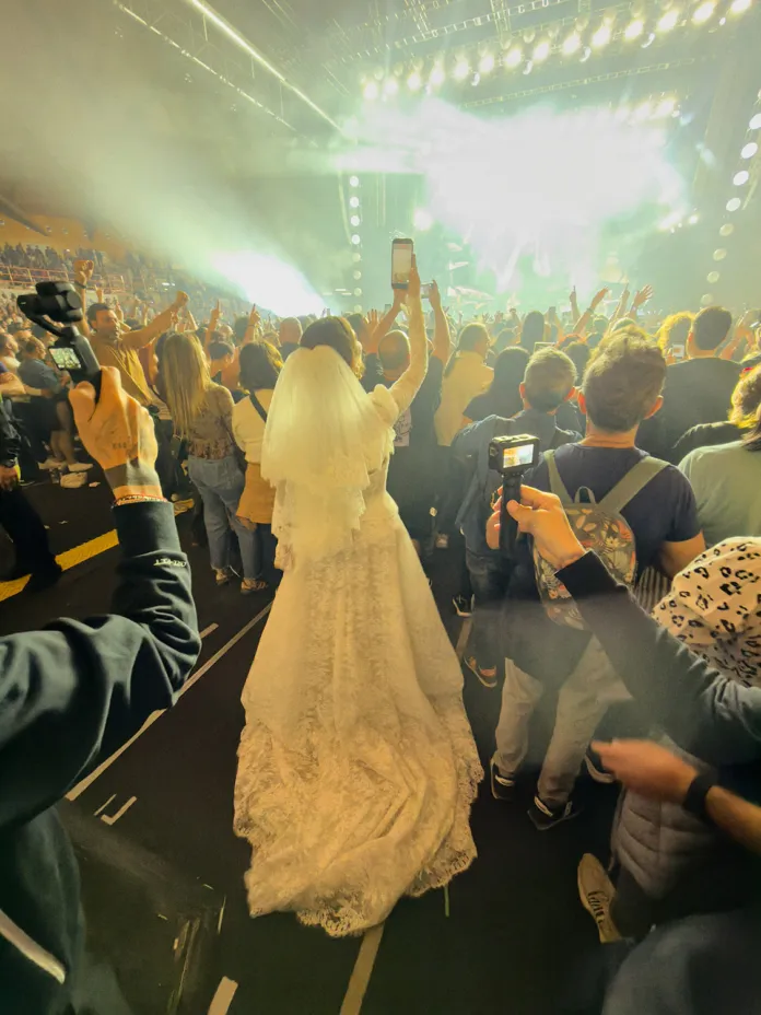 Immagine di una sposa che partecipa a un evento, vista di spalle con abito lungo e cappello, in un ambiente affollato con luci sul palco.
