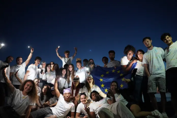 Un gruppo di giovani in maglietta chiara posa all’aperto di notte, tenendo una bandiera blu con stelle gialle.