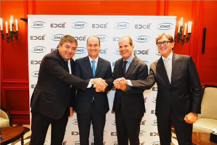 Quattro uomini in abiti formali posano davanti a un fondale ripetutamente decorato con i marchi CMD ed EDGE; due di loro si stringono la mano.
