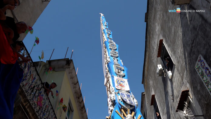 Foto di un giglio alto della festa di Barra osservato dal basso tra edifici con cielo azzurro.