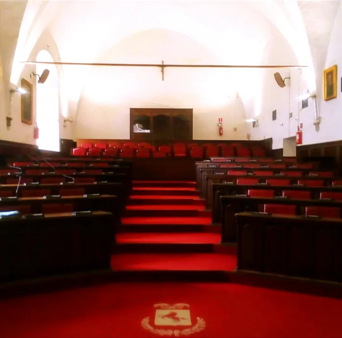 Immagine di archivio di una sala consiliare vuota con sedili in legno e tessuto rosso, presumibilmente di un'assemblea o consiglio.