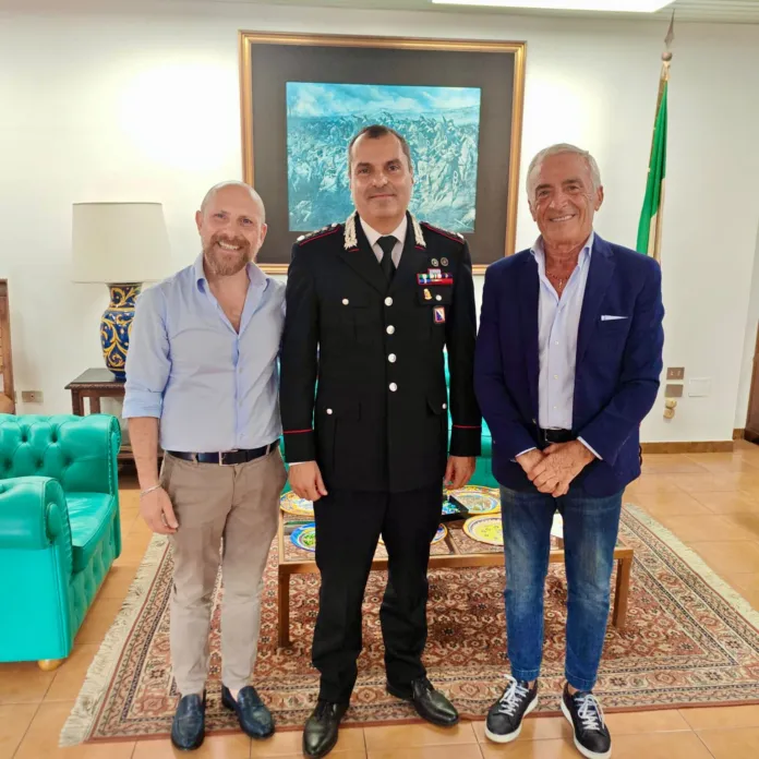 Tre uomini in piedi in un ufficio, uno in uniforme dei Carabinieri e due in abbigliamento civile elegante, in un contesto formale con arredamento classico.