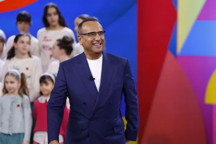 Un uomo in giacca blu sorride su un palco colorato; dietro di lui ci sono alcuni bambini.