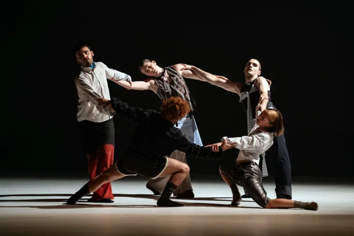 Un gruppo di danzatori si muove sul palcoscenico, illuminato su fondo nero.