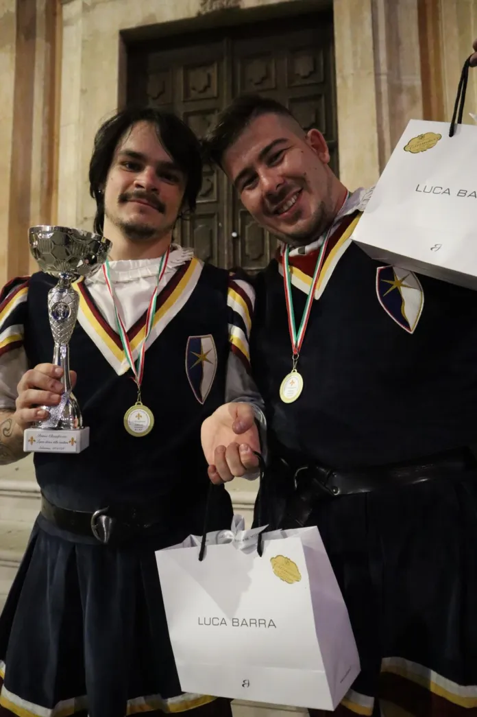 Due uomini in abiti scuri posano con medaglie al collo, un trofeo e sacchetti.