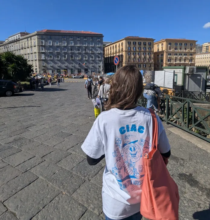 Una persona vista di spalle, con una maglietta recante la scritta CiAc, cammina su una piazza lastricata tra edifici e passanti.