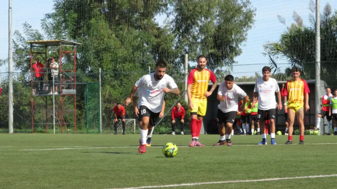 Un calciatore in maglia bianca è vicino al pallone; altri giocatori, con maglie bianche e giallorosse, si trovano alle sue spalle.