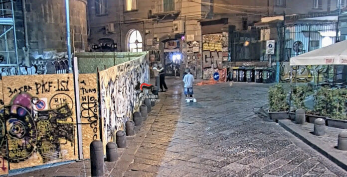 Piazza del Gesù a Napoli con muri coperti di graffiti e persone in lontananza in una strada di notte.